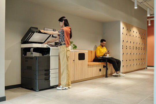 Young Asian woman using printer while young Black man sitting on bench with wooden lockers in background working on tablet in creative coworking space