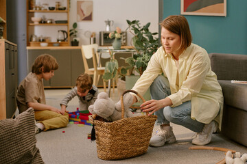 Caucasian middle aged woman organizing toys in basket, while two Caucasian children playing with building blocks on floor in living room during work from home situation