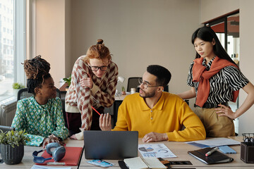 Fototapeta premium Young adult diverse men and women collaborating around laptop in modern coworking space, discussing project documents