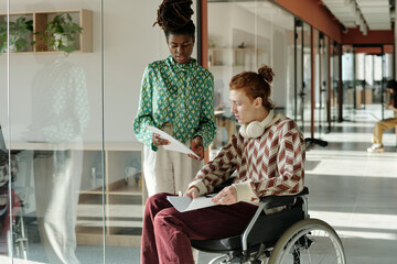 Young adult Black woman standing while discussing project with young adult Caucasian man in...