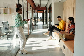 Young Black woman standing and gesturing while talking to young biracial man and young Asian woman sitting on bench in modern coworking office hallway