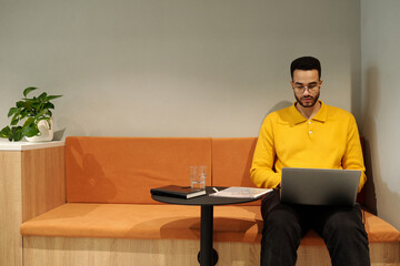 Young adult Black man wearing yellow sweater sitting on orange bench using laptop, reading...