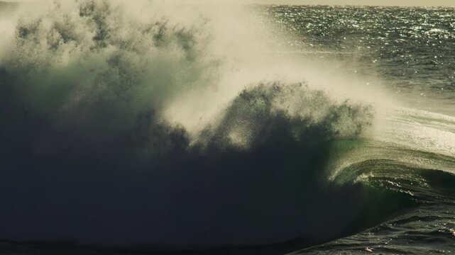 Dramatic wave builds in golden light as spray lifts off crest and deep shadows cut through sea