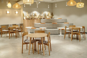 Modern coworking space featuring empty wooden tables and chairs, tiered seating with round cushions, neutral color palette, decorative dried plants