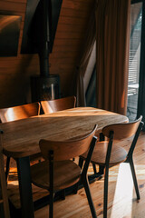 Wooden dining table with matching chairs in a cozy interior, illuminated by warm sunlight. A modern black wood stove stands in the background beside a large window with beige curtains