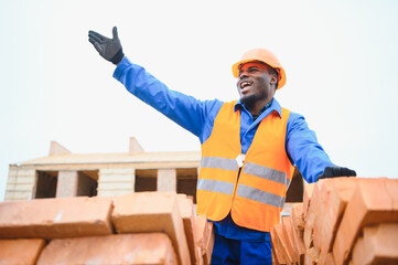 An African American construction worker, in work clothes and a hard hat, is building a brick house