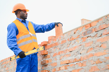 An African American construction worker, in work clothes and a hard hat, is building a brick house