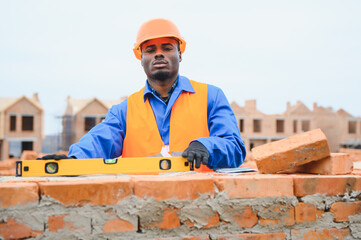 An African American construction worker, in work clothes and a hard hat, is building a brick house