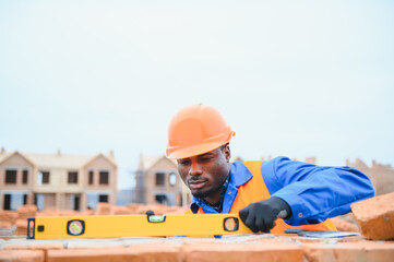 An African American construction worker, in work clothes and a hard hat, is building a brick house
