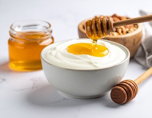Close up of a bowl of creamy yogurt topped with honey drizzled from a honey dipper, with a jar of honey and a bowl of nuts in the background on a white marble surface