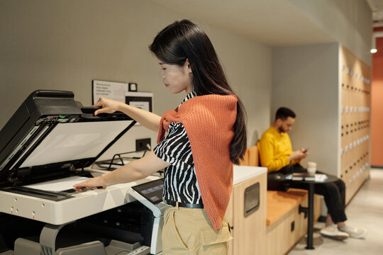 Asian woman scanning documents on office copier in modern coworking space while sitting man with beard using smartphone in background