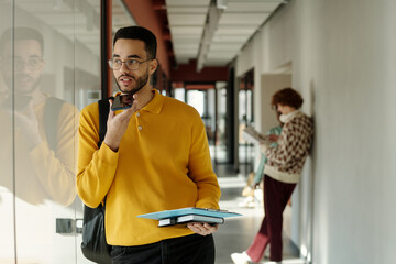 Young adult Biracial man wearing yellow sweater holding folders and using smartphone voice assistant while standing in modern coworking space