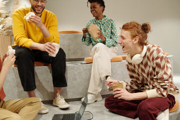 Group of young adult men and women of diverse ethnicities sitting on steps in creative coworking space, laughing and eating