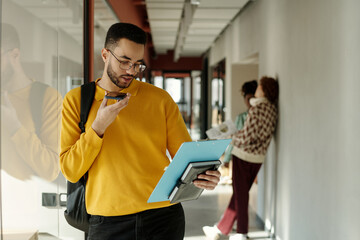 Biracial man with beard wearing yellow sweater holding clipboard and smartphone, recording voice...