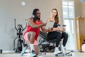 A man and a woman are in a gym. The man is showing the woman a tablet. The woman is holding two dumbbells