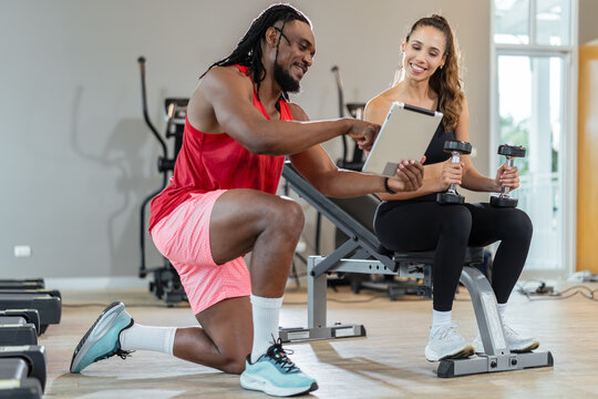 A man and a woman are in a gym. The man is pointing to a tablet. The woman is holding two dumbbells - Powered by Adobe