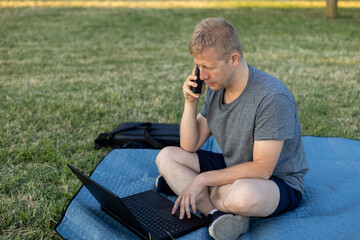 A middle-aged man sitting cross-legged on a blanket in a grassy park, holding a smartphone to his...