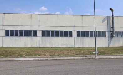 Concrete facade of factory building with row of window and a chimney. Meadow, sidewalk and road in...