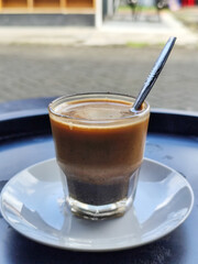 This close-up photo shows a cup of hot black coffee served on a small white plate with a spoon inside, placed on a black table in a roadside stall.