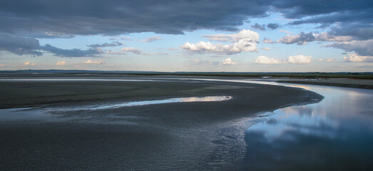 Calm waters reflect a dramatic sky at low tide near a tranquil estuary in the late afternoon