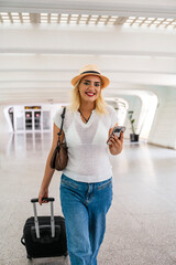 Young woman wearing hat and casual clothing walking in airport terminal while using smartphone and...