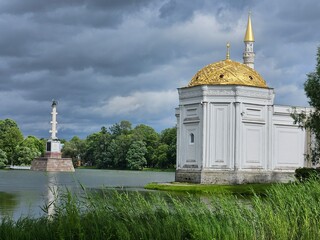 Large white building Turkish bath with gold accents sits next and Chesme column to a body of water....