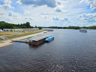 Naklejka premium Blue boat is docked next to a brown landing stage peer. The water is calm. There are several other boats in the water river Volkhov in Veliky Novgorod