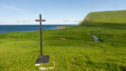 Black cross stands on lush green hillside overlooking serene ocean waves, with gentle slopes and a...