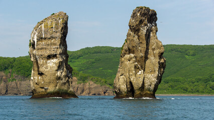 Two towering rock formations rise majestically from the calm waters, surrounded by lush green hills...