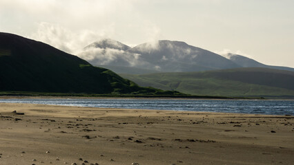 Serene landscape featuring rolling green hills and majestic mountains under a cloudy sky, with a calm river flowing along the sandy shore, creating a tranquil natural scene