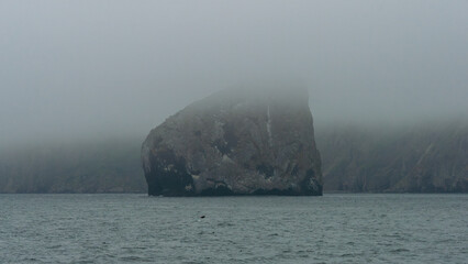 Large rock formation partially obscured by fog, rising from calm water, surrounded by misty cliffs,...