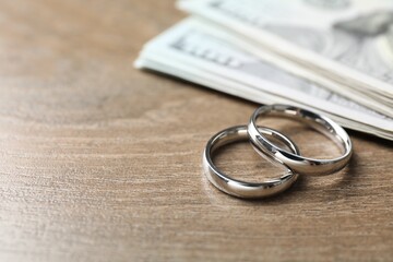 Silver wedding rings and dollar banknotes on wooden table, closeup. Space for text