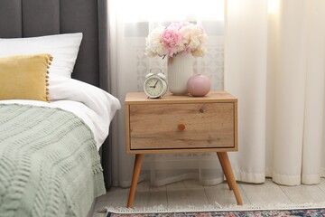 Bedside table with alarm clock and flowers near bed indoors