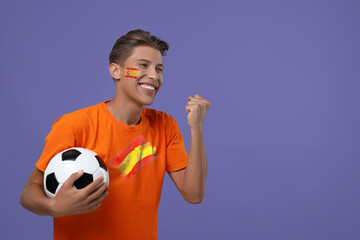Excited fan with flag of Spain on his face holding soccer ball against dark violet background....