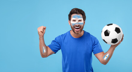 Excited fan with flags of Argentina painted on his arms holding soccer ball against light blue...