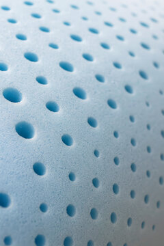 Close-up of perforated blue memory foam with evenly spaced round holes. The surface is soft, porous, and smooth with a subtle matte finish. The light falls diagonally