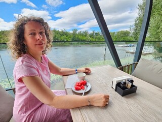 Woman is sitting at a table with a plate of fruit and a cup of coffee. She is wearing a pink dress...