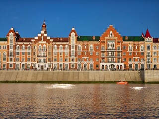 Row of buildings with a red roof and white trim. The buildings are on a river bank in Yoshkar-Ola