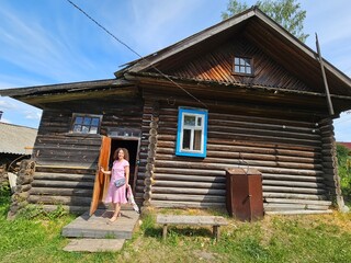 Woman in a pink dress stands in front of a wooden house. The house has a blue window and a brown...