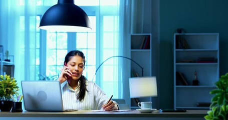 Businesswoman taking notes on phone call sitting at computer desk in modern cozy office, Indian Asian female using pencil to jot down pointers while talking on phone and working on laptop