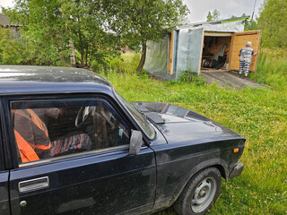 Black car is parked in a grassy area near garage with closing door. A person is sitting in the...
