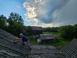 Man is on a roof, fixing something. The sky is cloudy. There are two houses in the background. The...