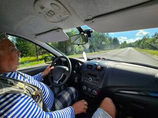 Man is driving a car with a blue stripe on his shirt. He is wearing plaid pants