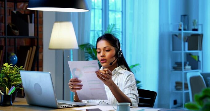 Businesswoman using headset for call at computer desk in modern cozy office, Indian Asian female attending teleconference with microphone headphones working on laptop communicating professionally