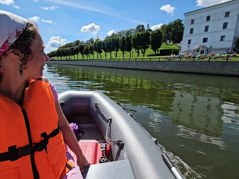 Woman in an orange life jacket is sitting in a inflatable boat on a river. The boat is small and has a red container on it near embankment