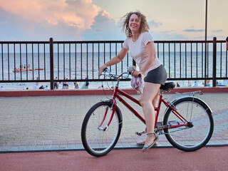 Woman is sitting on a red bicycle. She is wearing a pink shirt and shorts. The bicycle is parked on...