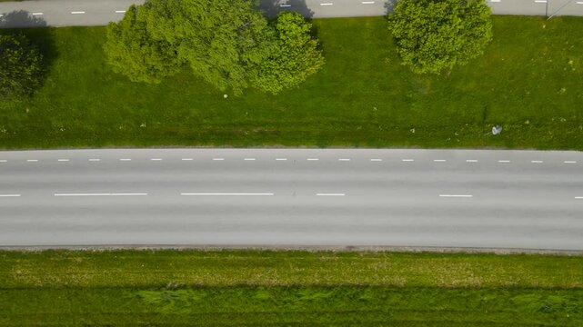 Top down aerial view of a rural and countryside paved or asphalt road where cars, vehicles and buses drive on back and fourth on a sunny summer day with green grass strip separating pedestrian road.