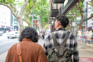 Two Chinese men in their early twenties, both wearing glasses, walk together along a wide and busy...