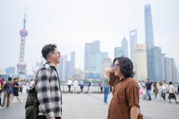 Two young Chinese men in their early twenties, both wearing glasses, face each other and talk along the riverside with tall skyscrapers in view across the water in May 2025 in Shanghai, China.