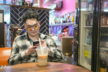 Two young Chinese men in their early twenties, both wearing glasses, are drinking beverages, using their smartphones, and relaxing at a cafe. Captured in May 2025 in North Bund, Shanghai, China.